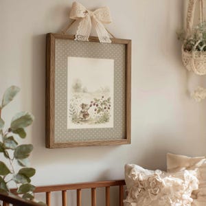 May include: A framed print featuring a watercolor illustration of a mouse gathering berries. The print is set within a wooden frame and adorned with a cream-colored lace bow. The background is a light gray with white polka dots.