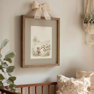 May include: A framed print featuring a watercolor illustration of a mouse gathering berries. The print is set within a wooden frame and adorned with a cream-colored lace bow. The background is a light gray with white polka dots.