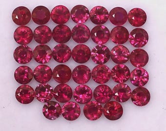 Puede incluir: Una colección de treinta y cuatro piedras preciosas redondas, facetadas y de color rojo vibrante dispuestas en una cuadrícula. Cada piedra presenta un corte brillante con múltiples facetas que reflejan la luz, creando un color carmesí profundo y uniforme.