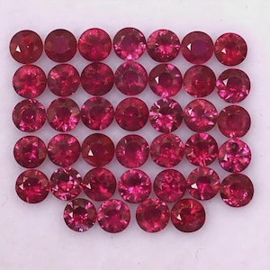 Puede incluir: Una colección de treinta y cuatro piedras preciosas redondas, facetadas y de color rojo vibrante dispuestas en una cuadrícula. Cada piedra presenta un corte brillante con múltiples facetas que reflejan la luz, creando un color carmesí profundo y uniforme.