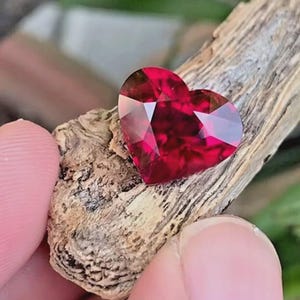 Puede incluir: Una piedra preciosa en forma de coraz&oacute;n de color rojo intenso con m&uacute;ltiples facetas que reflejan la luz. La piedra est&aacute; colocada sobre un trozo de madera de color marr&oacute;n claro texturizado con patrones de veta visibles y bordes rugosos.