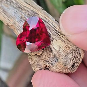 Puede incluir: Una piedra preciosa facetada en forma de coraz&oacute;n de color rojo intenso con un corte brillante. La piedra est&aacute; colocada sobre un trozo de madera texturizada de color marr&oacute;n claro, mostrando su superficie reflectante y bordes sim&eacute;tricos.