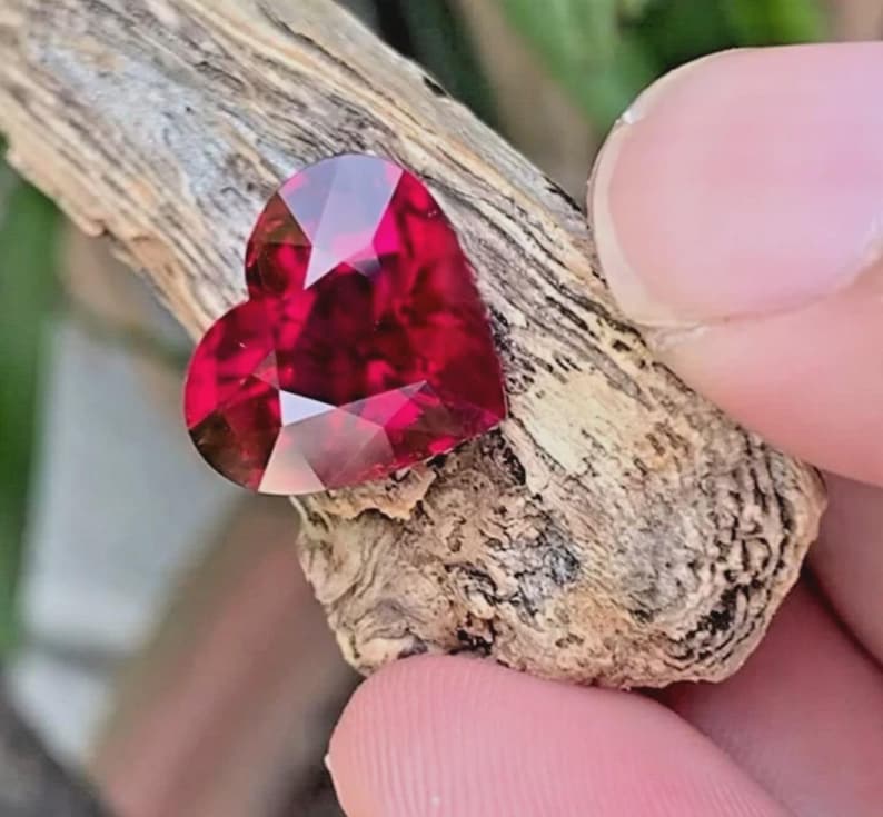 Puede incluir: Una piedra preciosa facetada en forma de coraz&oacute;n de color rojo intenso con un corte brillante. La piedra est&aacute; colocada sobre un trozo de madera texturizada de color marr&oacute;n claro, mostrando su superficie reflectante y bordes sim&eacute;tricos.