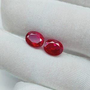 Può includere: Due rubini rosso vivo dal taglio ovale con superfici sfaccettate che riflettono la luce. Le pietre preziose sono posizionate fianco a fianco, mostrando il loro profondo colore cremisi e la loro chiarezza lucida. Ogni rubino è simmetrico.