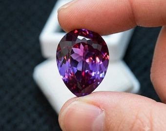 Puede incluir: Una piedra preciosa facetada en forma de pera que muestra un degradado de color vibrante que transita de púrpura intenso a magenta y rosa brillante. La piedra presenta un corte brillante con múltiples superficies reflectantes.