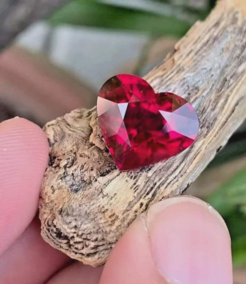 Puede incluir: Una piedra preciosa en forma de coraz&oacute;n de color rojo intenso con m&uacute;ltiples facetas que reflejan la luz. La piedra est&aacute; colocada sobre un trozo de madera de color marr&oacute;n claro texturizado con patrones de veta visibles y bordes rugosos.
