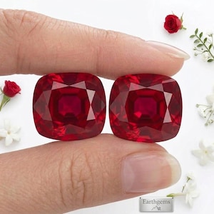 May include: Two cushion-cut, deep red gemstones with faceted surfaces. The stones are held between fingers against a white background decorated with small red roses and white flowers. A small silver rectangular tag displays the text Earthgems.