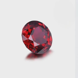 May include: A round, faceted gemstone with a deep, vibrant red colour. The stone features a brilliant cut with multiple triangular and kite-shaped facets that reflect light, creating a complex pattern of dark and light red tones across its surface.