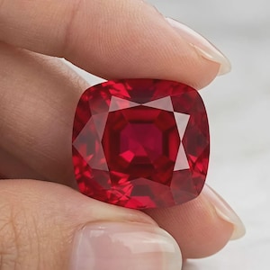 May include: A cushion-cut gemstone with a deep, vibrant red color and a faceted surface that reflects light. The stone has a square shape with rounded corners and exhibits high clarity, showcasing a rich, saturated crimson hue throughout its body.