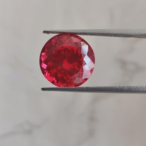 Könnte beinhalten: Ein runder, facettierter, leuchtend roter Edelstein, der von einer silberfarbenen Metallpinzette gehalten wird. Der Stein weist einen Brillantschliff mit zahlreichen dreieckigen und rautenförmigen Facetten auf, die das Licht reflektieren.