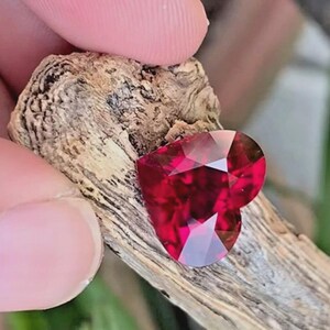 Puede incluir: Una piedra preciosa facetada en forma de coraz&oacute;n de color rojo intenso con un corte brillante, apoyada contra un trozo de madera desgastada de color marr&oacute;n claro y textura rugosa. La piedra muestra facetas geom&eacute;tricas n&iacute;tidas.