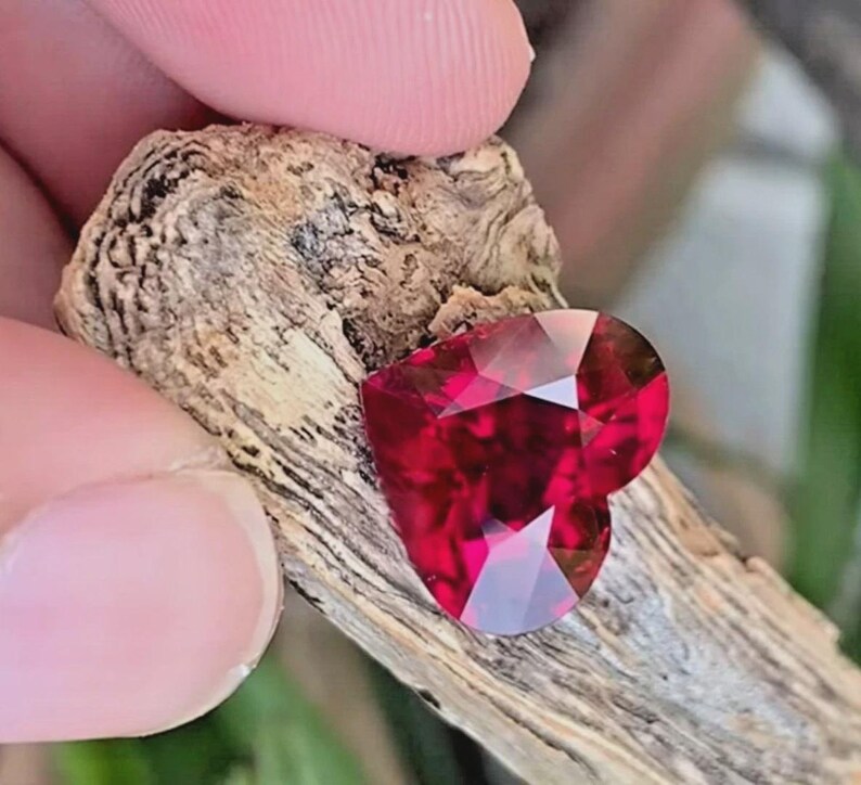 Puede incluir: Una piedra preciosa facetada en forma de coraz&oacute;n de color rojo intenso con un corte brillante, apoyada contra un trozo de madera desgastada de color marr&oacute;n claro y textura rugosa. La piedra muestra facetas geom&eacute;tricas n&iacute;tidas.