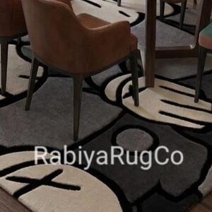 May include: A rectangular area rug featuring a repeating graphic pattern of stylized cartoon hands and faces with X-shaped eyes in shades of gray, black, and cream. The text RabiyaRugCo is printed in white across the center of the rug surface.