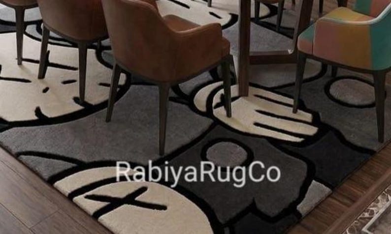 May include: A rectangular area rug featuring a repeating graphic pattern of stylized cartoon hands and faces with X-shaped eyes in shades of gray, black, and cream. The text RabiyaRugCo is printed in white across the center of the rug surface.