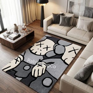 May include: A rectangular area rug featuring a stylized character design with X-shaped eyes in shades of cream, light gray, and dark charcoal gray. The rug displays the text "RabiyaRugCo" in white lettering across the center of the graphic pattern.
