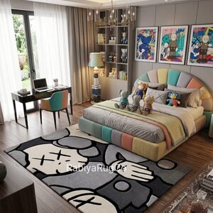 May include: A bedroom featuring a large area rug with a gray, black, and white cartoon character graphic and the text "RabiyaRugCo". The room includes a bed with a multi-colored headboard, several plush toys, a desk, a chair, and wall art.