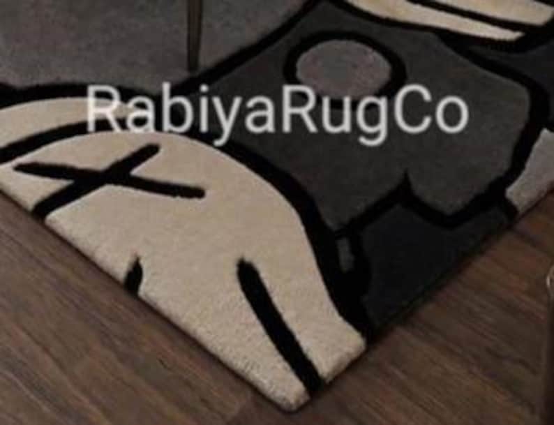 May include: A tufted area rug featuring a graphic design in cream, dark gray, and black. The rug displays bold black outlines and the text "RabiyaRugCo" printed in white across the center. The surface has a thick, textured pile construction.
