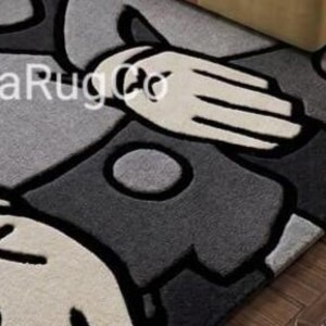 May include: A rectangular area rug featuring a stylized graphic design in shades of black, dark gray, and light gray. The pattern includes large, rounded shapes and hand-like forms outlined in thick black lines. The text biyaRugCo is visible.