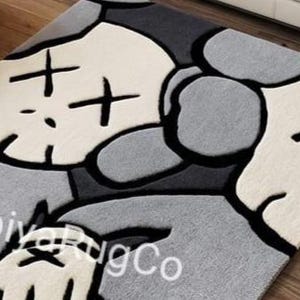 May include: A rectangular area rug featuring a stylized character graphic in shades of cream, light gray, dark gray, and black. The design includes a large head with X-shaped eyes, rounded ears, and a gloved hand. The text abiyaRugCo is visible.