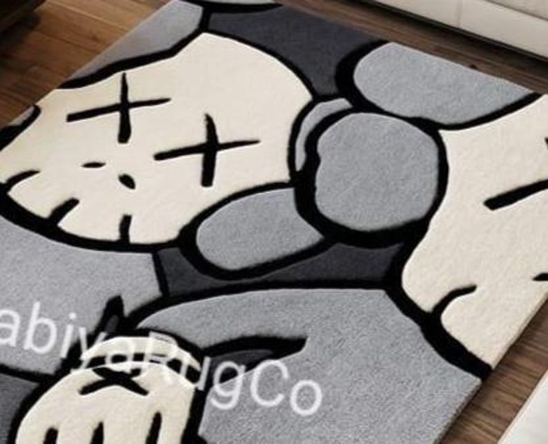 May include: A rectangular area rug featuring a stylized character graphic in shades of cream, light gray, dark gray, and black. The design includes a large head with X-shaped eyes, rounded ears, and a gloved hand. The text abiyaRugCo is visible.