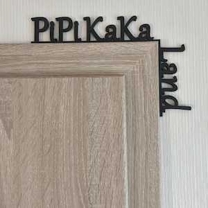 Könnte beinhalten: Schwarzes Holzschild mit dem Text "PiPi Kaka Land" an der Ecke einer hellbraunen Holztür angebracht. Das Schild ist an einer weißen Wand befestigt. Die Tür hat einen silbernen Griff.