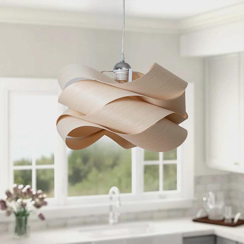 May include: A modern, light-colored wooden pendant light fixture. The fixture features a layered, wavy design, suspended from a silver-toned ceiling mount. The light hangs in front of a window, illuminating a kitchen space.