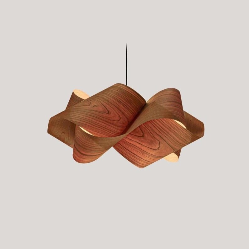 May include: A modern, sculptural pendant light fixture. The light features a unique design with layered, curved wooden panels, showcasing a natural wood grain pattern. The fixture hangs from a black cord, creating a warm, ambient glow.