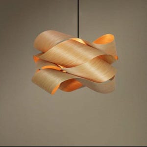 May include: A modern pendant light fixture with a unique design. The light is made of layered, curved wood in a light brown color. The interior of the light fixture is a warm orange color, creating a soft glow. The light hangs from a black cord.