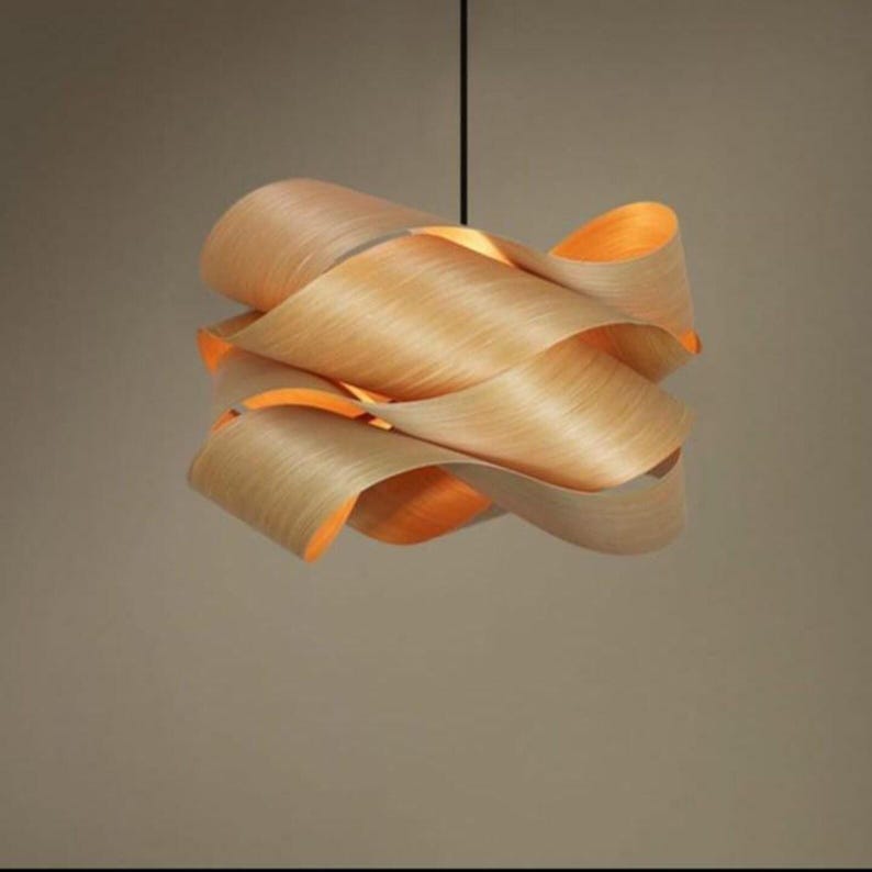 May include: A modern pendant light fixture with a unique design. The light is made of layered, curved wood in a light brown color. The interior of the light fixture is a warm orange color, creating a soft glow. The light hangs from a black cord.