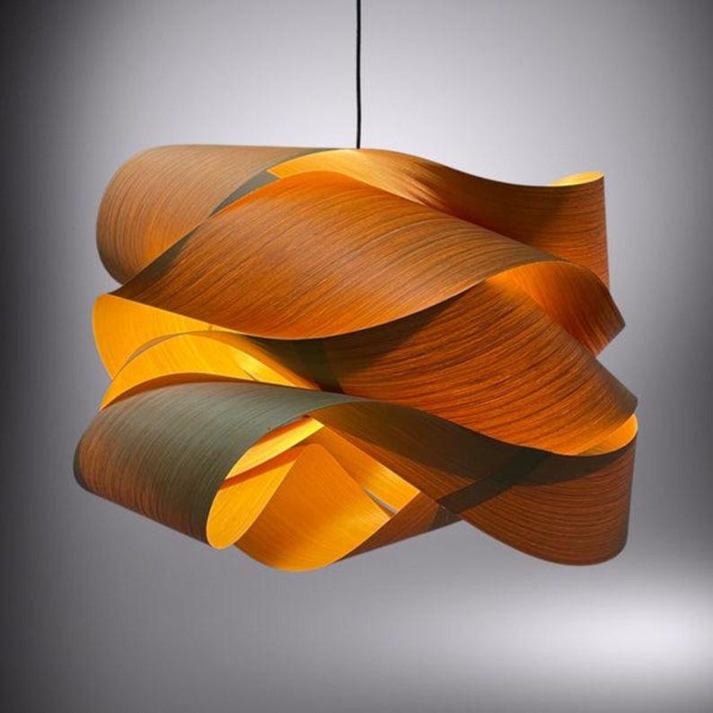May include: A modern pendant light fixture with a unique design. The light is made of layered, curved wooden panels in shades of brown and orange. The fixture is suspended from a black cord and emits a warm, inviting glow.