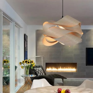 May include: A modern, sculptural pendant light fixture with a layered, wave-like design. The light is made of a light-colored material, possibly wood, and hangs from a black cord. The fixture is the focal point of a contemporary living room.