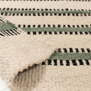 Puede incluir: Una alfombra tejida con una base texturizada de color blanco roto y rayas horizontales en verde salvia apagado y negro. La alfombra tiene una construcción de pelo rizado grueso y está doblada en una esquina para mostrar el reverso.