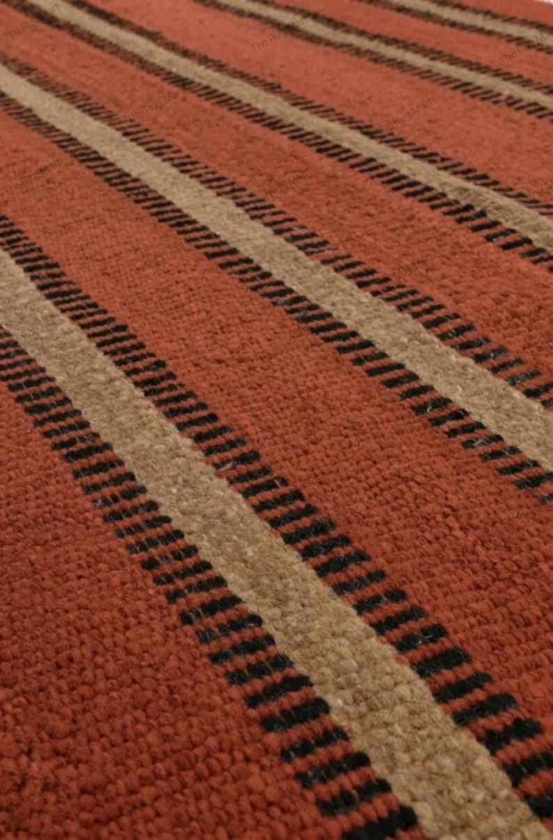 May include: A woven rug featuring a repeating pattern of wide terracotta orange stripes alternating with narrower beige stripes, each bordered by thin, dark black lines. The texture appears dense and fibrous with a consistent, uniform pile.