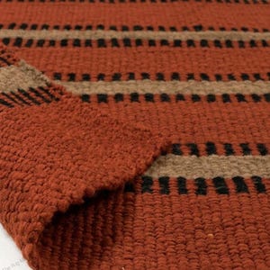 May include: A woven area rug featuring a textured, chunky knit surface in a deep terracotta orange color. The rug displays a repeating horizontal pattern of wide terracotta bands, narrower tan stripes, and thin, dashed black lines throughout.