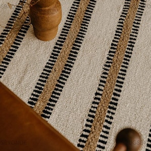 May include: A woven rug with a cream-colored background and vertical stripes in black and tan. A brown vase with dried branches sits on the rug. The rug is likely made of natural fibers.
