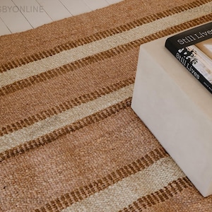 May include: A striped rug in shades of beige and brown, with a textured weave. The rug is laid on a light wooden floor. A white, square side table is in the background, with a book titled "Still Lives" resting on it.