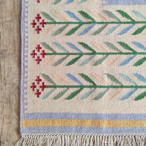 Puede incluir: Una alfombra tejida con un fondo color crema y un patrón repetido de hojas verdes y azules. La alfombra tiene detalles florales rojos y un borde azul claro. El borde inferior presenta flecos.
