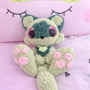 May include: A handmade, green and teal crochet frog plushie with pink accents on its paws and large, glittery eyes. The plushie is displayed on a pink pillow with the text "PDF pattern" on a banner.