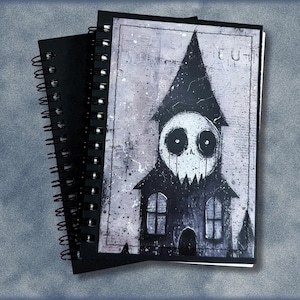 Puede incluir: Un cuaderno de espiral con una ilustración gráfica en blanco y negro de una casa con un rostro grande, redondo y similar a una calavera integrado en el techo. La obra tiene un aspecto texturizado y desgastado con salpicaduras y sombras.