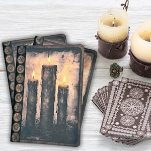 May include: A stack of tarot cards with a brown and white geometric design. Three lit candles with dripping wax sit near the cards. A green jeweled ring and two candles in brown holders are also present. The cards feature an image of three lit candles.