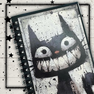 May include: A spiral-bound notebook featuring a black and white graphic illustration of a stylized cat with large round eyes, a wide toothy grin, and a small red nose. The cover design includes splattered paint effects and abstract text.