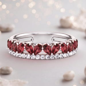 Op de afbeelding: Een zilverkleurige ring met een rij dieprode, peervormige edelstenen in een hartvormig patroon, geplaatst boven een enkele rij kleine, heldere, rondgeslepen stenen die de ring omcirkelen in een klassiek eternity-ontwerp.