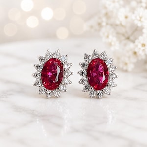 May include: A pair of oval-shaped ruby earrings with a halo of small, sparkling white gemstones. The earrings are set in a silver metal and rest on a white marble surface. The background is blurred with soft, out-of-focus lights.
