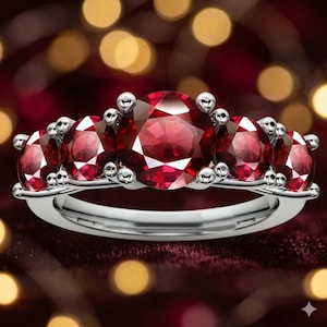 May include: A silver ring featuring five large, round, vibrant red gemstones. The gemstones are set in a row, with smaller silver accents between each stone. The ring is displayed against a blurred background of red and gold.
