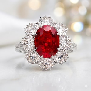 May include: A silver ring featuring a large, oval-cut ruby surrounded by a halo of smaller, round-cut diamonds. The ring's band is also adorned with smaller diamonds, creating a sparkling, elegant design. The ruby is a deep, vibrant red.