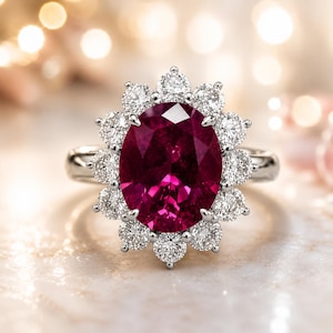 Op de afbeelding: Een zilveren ring met een grote, ovaal geslepen robijn omringd door een halo van kleinere, ronde diamanten. De robijn is dieprood en de diamanten schitteren. De ring ligt op een wit marmeren oppervlak.