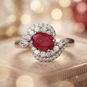 Op de afbeelding: Een ring met een centrale ovaal geslepen dieprode robijn. De robijn is omringd door een wervelende, asymmetrische halo van kleine, ronde, briljant geslepen witte diamanten, gezet in een gepolijste metalen band die om de steen heen krult.
