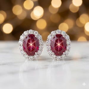 May include: A pair of oval-shaped ruby earrings with a halo of small, round, clear gemstones. The rubies have a deep red color and are set in a silver or white metal. The background is blurred with golden bokeh lights.