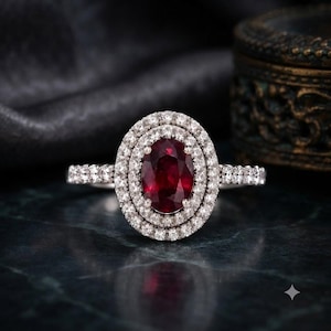 Op de afbeelding: Een zilverkleurige ring met een centrale ovaal geslepen dieprode edelsteen, omringd door een dubbele halo van kleine, heldere, rond geslepen stenen. De ringband is aan beide zijden bezet met een rij kleine, heldere, rond geslepen stenen.
