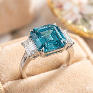 Könnte beinhalten: Ein silberner Ring mit einem großen, quadratischen, teal-farbenen Edelstein als Mittelpunkt, flankiert von zwei kleineren, rechteckigen, klaren Edelsteinen. Der Ring ist in einem klassischen, eleganten Stil gehalten, ideal für besondere Anlässe.